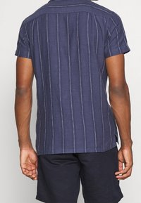 Camicia a maniche corte in tessuto blu scuro con righe bianche verticali, dotata di colletto e vestibilità comoda. Vista posteriore, pensata per il comfort.