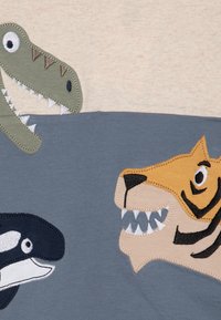 Et lys beige stoff har lekne dyre-applikasjoner: en grønn dinosaur, en spekkhogger og en brun tiger, hver med broderte detaljer og teksturer.