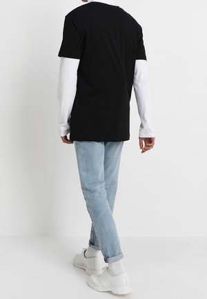 Person trägt ein schwarzes Kurzarmshirt über einem weißen Langarmshirt, hellblaue Jeans, weiße Socken und weiße Schuhe, steht an einer weißen Wand.