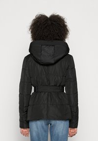 Sort pufferjakke med tætsiddende talje, hætte og elastiske detaljer. Har quiltede mønstre og en glat, skinnende tekstur.