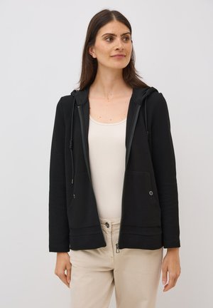 Zwarte zip-up hoodie van een gestructureerde stof, met een capuchon met koordjes en twee voorzakken; gedragen over een licht tanktop en beige broeken.
