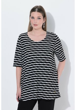 Frau trägt ein kurzärmeliges Oberteil mit schwarz-weißen, wellenförmigen Streifen und silberne, hängende Ohrringe, steht vor einem schlichten hellen Hintergrund.