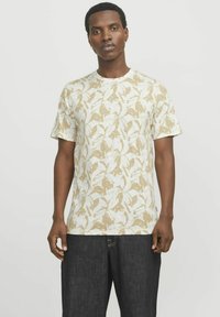 Camiseta de manga corta, color beige claro con patrones de hojas blancas, escote redondo, corte casual, hecha de tela texturizada. Combinada con jeans oscuros.