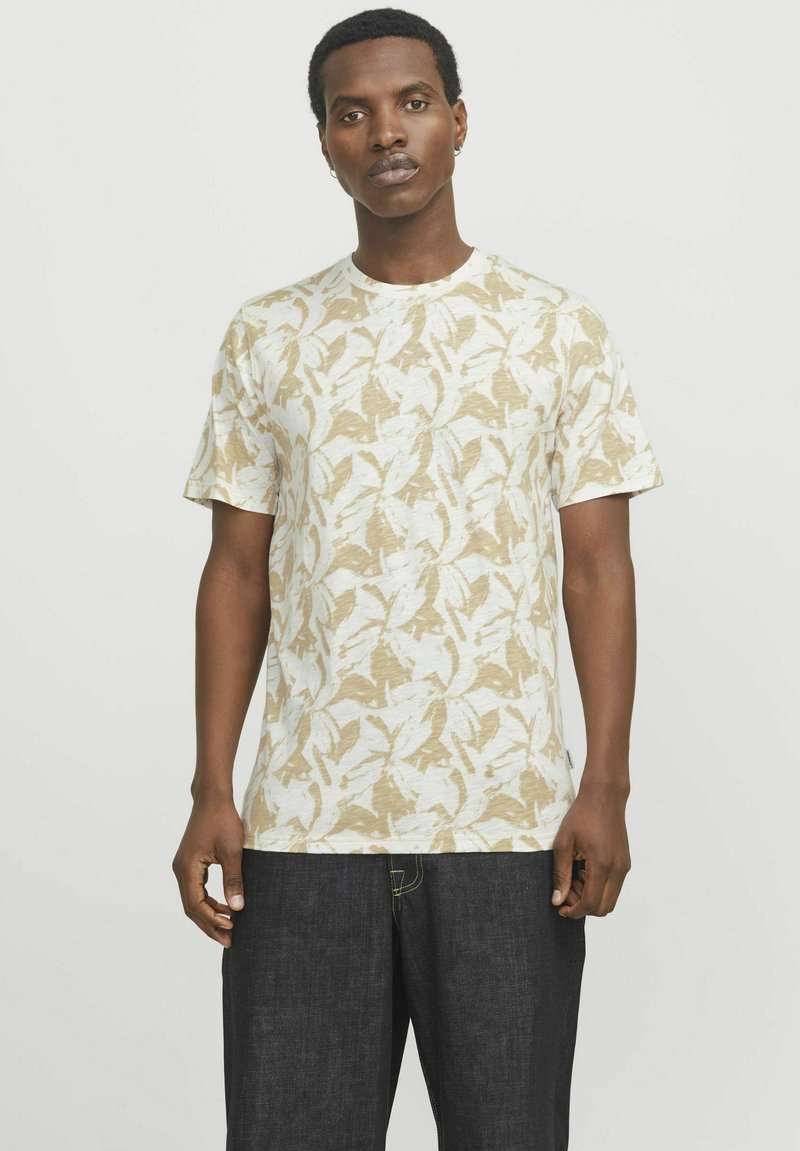 T-shirt a maniche corte, beige chiaro con motivi a foglia bianchi, scollo rotondo, vestibilità casual, realizzato in tessuto testurizzato. Indossato con jeans scuri.