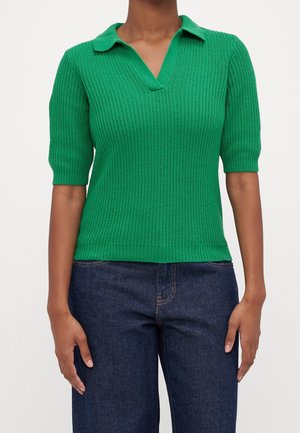 Personne portant un polo en maille côtelée vert à manches courtes et un jean bleu foncé, debout devant un fond blanc uni.