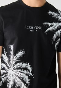 T-shirt en coton noir avec des graphiques de palmiers blancs et le texte "PIER ONE BERLIN", col rond, manches courtes et texture lisse.