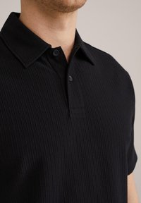 Črna rebrasta polo majica s ovratnikom, kratkimi rokavi in dvema gumboma na zapenjanju. Narejena iz strukturiranega materiala, ki prikazuje subtilne navpične črte.