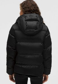 Chaqueta acolchada negra con un acabado texturizado y capucha. Presenta un diseño ajustado con bafles horizontales y un logo en la parte de atrás.