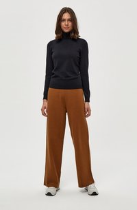 Dunkel gestrickter Rollkragenpullover kombiniert mit weit geschnittenen braunen Hosen; das Model trägt weiße Sneaker. Glatte Texturen, neutrales Farbschema.
