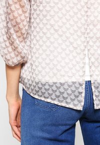 Chemisier beige transparent avec un motif géométrique, texture douce et manches bouffantes, associé à un jean en denim bleu.