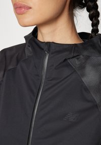 Schwarze Sportjacke mit Reißverschluss, strukturierten Akzenten und dezentem Logo. Der Stoff ist leicht und hat eine glatte Oberfläche.