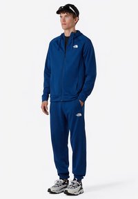 Modrá sportovní souprava se zipovou bundou s kapucí a odpovídajícími kalhotami. Vyznačuje se měkkou texturou, volným střihem a bílými logy jako doplňky.