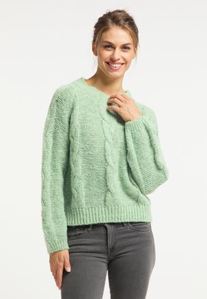 Lachende vrouw met een lichtgroene kabelgebreide trui en donkergrijze spijkerbroek tegen een effen witte achtergrond.