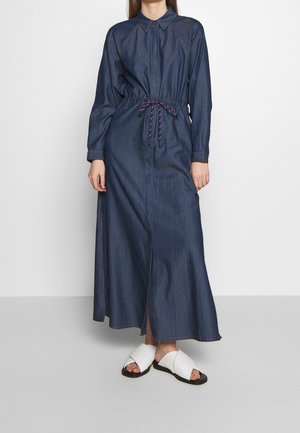 Kobieta w długiej sukience maxi z ciemnoniebieskiego denimu z długim rękawem i talią podkreśloną sznurkiem, ubrana w białe sandały z krzyżującymi się paskami, stoi na jednolitym tle.