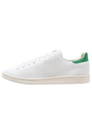 Biały niski sneaker o dzianej fakturze, z zielonym paskiem na pięcie, białymi sznurowadłami i beżową podeszwą z wytłoczonym logo Adidas, widok z boku.