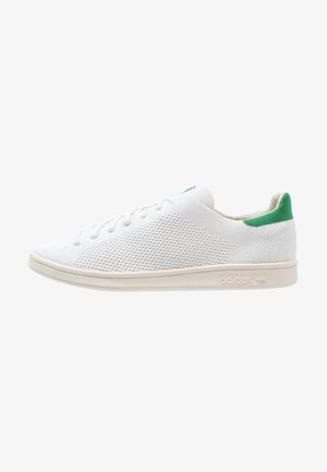 Biały niski sneaker o dzianej fakturze, z zielonym paskiem na pięcie, białymi sznurowadłami i beżową podeszwą z wytłoczonym logo Adidas, widok z boku.