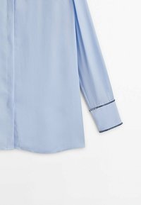 Chemise bleu clair à manches longues avec une texture lisse, patte de boutonnage sur le devant et décoration le long des poignets, présentant une coupe droite.