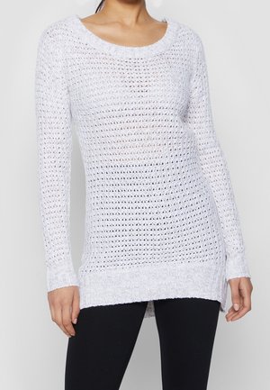 Femme portant un pull blanc à manches longues en maille ajourée avec un col rond et un pantalon noir sur un fond uni.