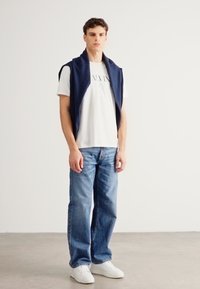 Chaleco de punto azul marino sobre una camiseta blanca con texto, combinado con vaqueros azules y zapatillas blancas, en un entorno luminoso y minimalista.