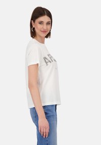 Weißes Baumwoll-T-Shirt mit kurzen Ärmeln, das mit silbernen Perlenbuchstaben "AFR" über der Brust verziert ist. Kombiniert mit blauen Denim-Jeans.