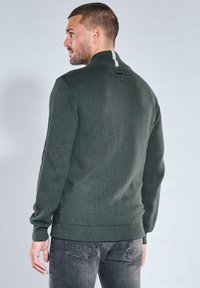 Dunkelgrüner, gestrickter Pullover mit hohem Kragen, mit einem strukturierten Muster und weißem Streifenakzent auf der Rückseite. Taillierte Passform mit gerippten Bündchen.