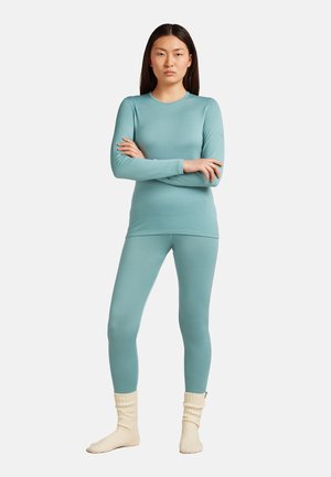 Woman with long dark hair stands with arms crossed wearing light blue long-sleeve top and leggings, and cream-colored knitted socks.