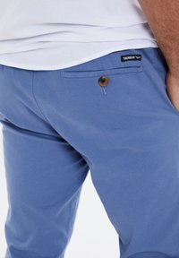 Chino in cotone blu con una texture liscia, dotato di un dettaglio in plastica marrone e una tasca posteriore. Logo visibile sulla vita.