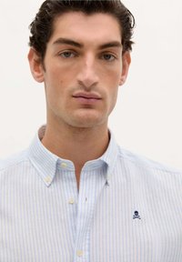 Camicia a maniche lunghe a righe blu chiaro con colletto, caratterizzata da un piccolo logo ricamato di teschio e ossa incrociate sul petto. Tessuto dalla consistenza morbida.