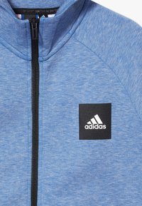 Modrá sportovní bunda s vysokým límcem, plným zipem vpředu a černou nášivkou s logem adidas na levé hrudi. Měkká, texturovaná látka.