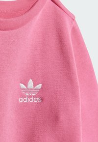 Rosa hettegenser med strukturert stoff, rund hals og en hvit brodert Adidas-logo med tre blader.