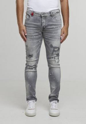 Homme portant un jean gris usé avec des éclaboussures de peinture blanche, des baskets blanches et un débardeur blanc, debout devant un fond uni.