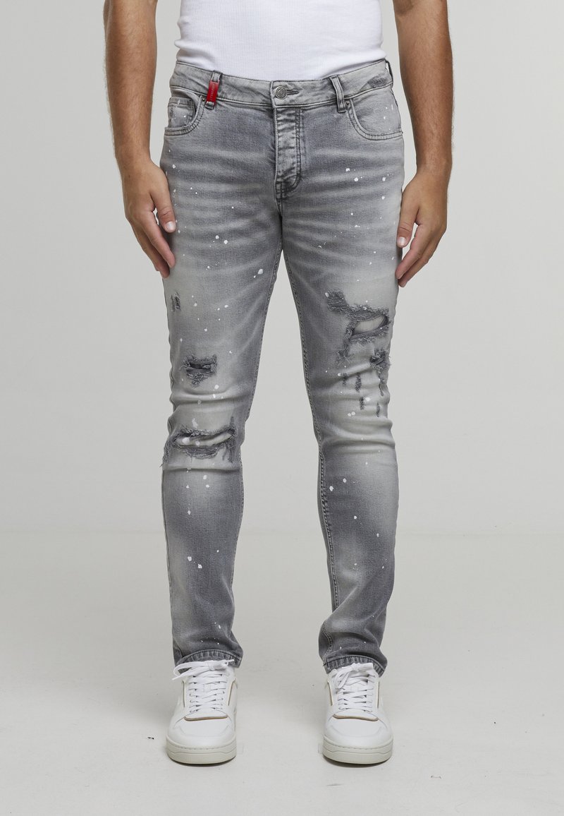 Homme portant un jean gris usé avec des éclaboussures de peinture blanche, des baskets blanches et un débardeur blanc, debout devant un fond uni.
