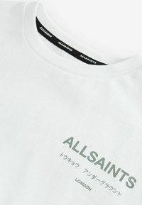 Biela krátka pánska tričko s krátkymi rukávmi vyrobené z bavlny, s okrúhlym výstrihom. Obsahuje zelený nápis "ALLSAINTS" a dodatočný japonský a anglický text nižšie.