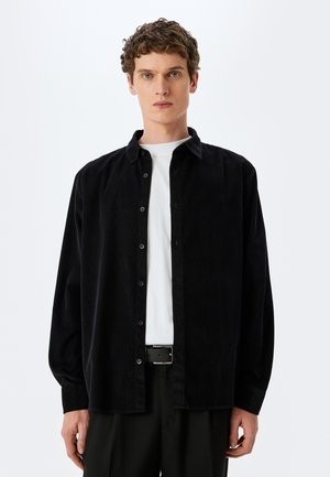 Camicia di velluto nero con chiusura a bottoni, maniche lunghe e colletto, indossata sopra una t-shirt bianca, abbinata a pantaloni scuri e una cintura.