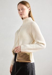 Biały dziany sweter z golfem typu mock z fakturą prążkowaną, zestawiony z czarną spódnicą i błyszącą złotą, prostokątną torebką na ramię.