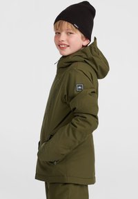 Veste isolée verte avec capuche, dotée de poches zippées et d'un patch logo sur la manche. Le modèle porte un bonnet en tricot noir.