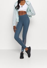 Kvinna klädd i vit Adidas sport-bh, ljusblå jacka, blå högmidjade leggings och svarta Adidas-sneakers stående med korsade ben.