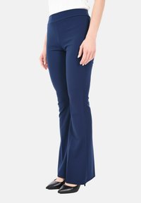 Donna che indossa pantaloni a gamba svasata blu navy aderenti e décolleté neri con punta affusolata su uno sfondo bianco.