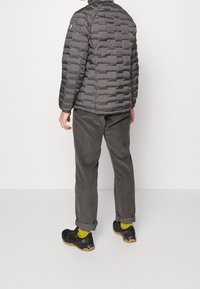 Giacca grigia imbottita con baffi orizzontali, abbinata a pantaloni in velluto a coste grigio scuro e scarpe nere. Calzini verde brillante visibili.