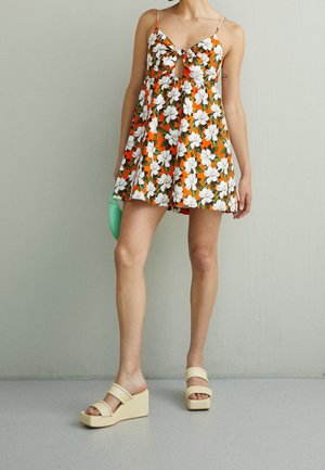 Žena v oranžových květovaných minišatech s tenkými ramínky a krémovými klínovými sandály stojí u světle šedé zdi.