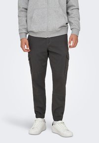 Sudadera con capucha y cremallera gris sobre pantalones cargo oscuros, con bolsillos laterales, puños elásticos y zapatillas blancas, sobre un fondo liso.