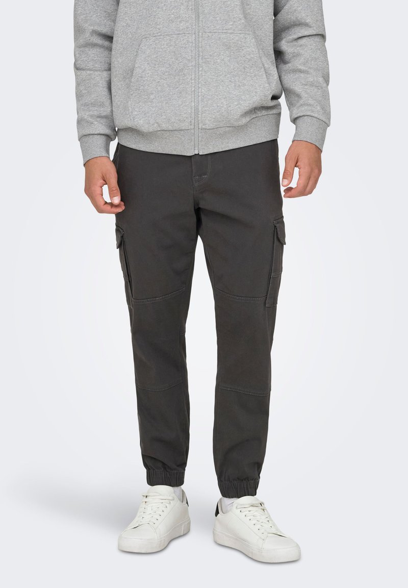 Sudadera con capucha y cremallera gris sobre pantalones cargo oscuros, con bolsillos laterales, puños elásticos y zapatillas blancas, sobre un fondo liso.