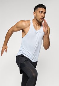 Helle graue sportliche Tanktop mit strukturiertem Finish, kombiniert mit schwarzen enganliegenden Shorts. Das Model befindet sich in einer Laufpose.