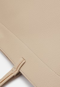 Gros plan sur l'angle d'un sac à main en cuir beige texturé montrant les coutures et la fixation de la poignée.