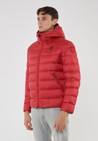 Giovane uomo che indossa una giacca a cappuccio invernale rossa, trapuntata, con tasche con zip e jeans chiari, in piedi davanti a uno sfondo semplice.