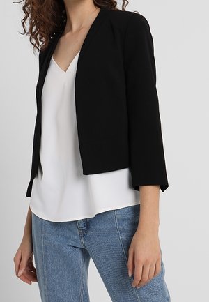 Frau trägt einen schwarzen kurz geschnittenen Blazer über einer weißen Bluse mit V-Ausschnitt und hellblauen Jeans, steht vor einem schlichten Hintergrund.