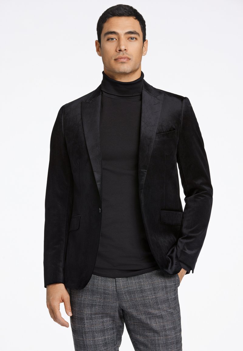 Zwarte fluwelen blazer met satijnen revers en een enkele zak, gecombineerd met een fitted zwarte coltrui, gedragen over een geruite broek.