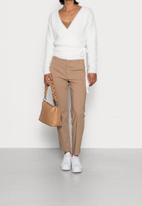 Witte v-hals wikkeltrui met een strik, beige aangesloten broek en een lichtbruine handtas met een geplooid handvat; witte sneakers maken de look compleet.