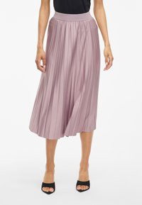 Záhybová midi sukňa v jemnej levanduľovej tkanine, s širokým elastickým pásom a splývavým siluetom. Nosí sa s čiernymi lodičkami s otvorenou špičkou.