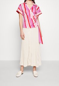 Blouse portefeuille rayée aux nuances roses et rouges, à manches courtes et nouée à la taille, associée à une jupe crème plissée à boutons.